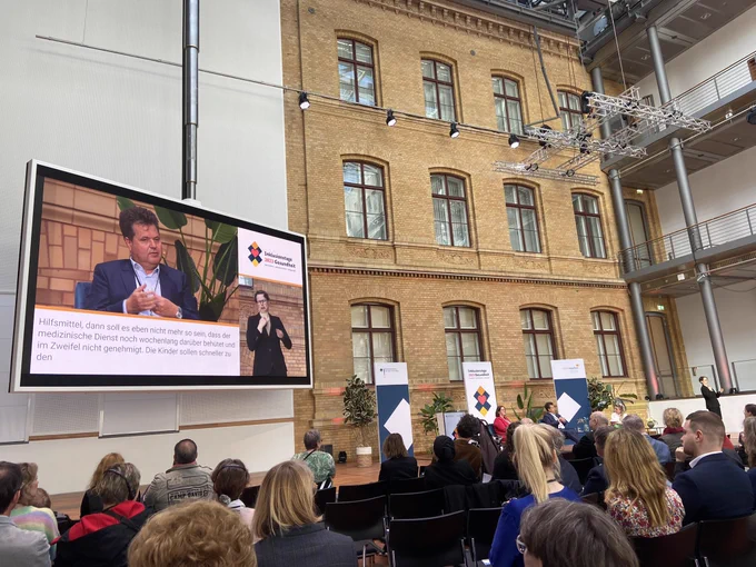Publikum und Podium mit Jürgen Dusel, Beauftragter der Bundesregierung für die Belange von Menschen mit Behinderung auf der Leinwand und Kerstin Griese MdB, SPD, mit der Moderatorin Nina LaGrande in einem großen Raum man sieht ein Haus