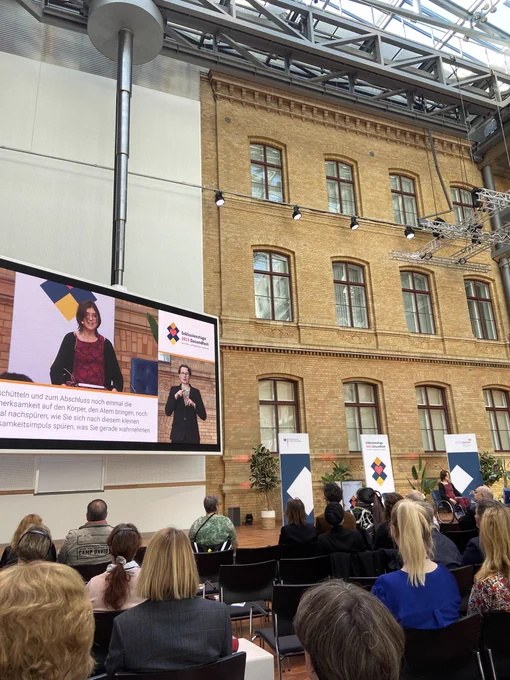 Publikum und Podium mit Jürgen Dusel, Beauftragter der Bundesregierung für die Belange von Menschen mit Behinderung und Kerstin Griese MdB, SPD, auf der Leinwand mit der Moderatorin Nina LaGrande in einem großen Raum man sieht ein Haus