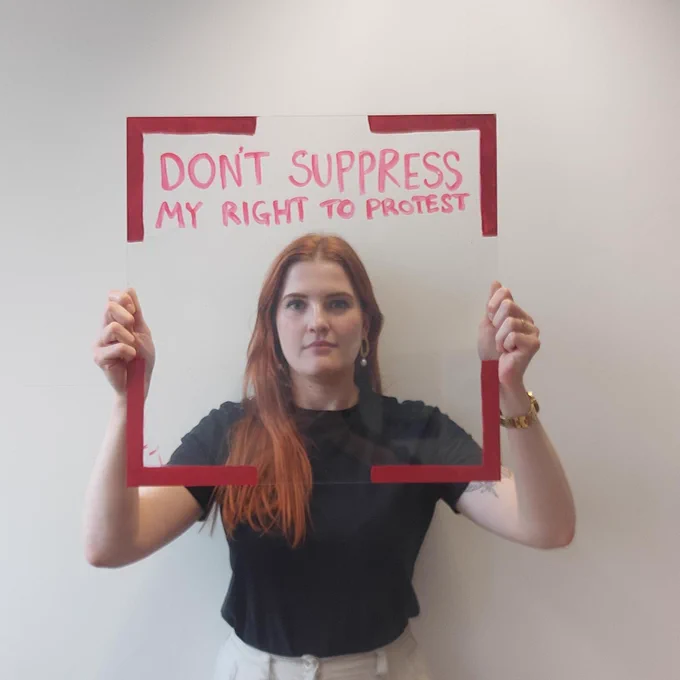 A woman holds a frame with red egdes and the words 'don't suppress my right to protest'