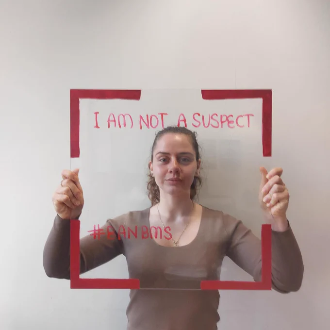 A woman holds a frame with red egdes and the words 'I am not a suspect' and 'BanBMS'