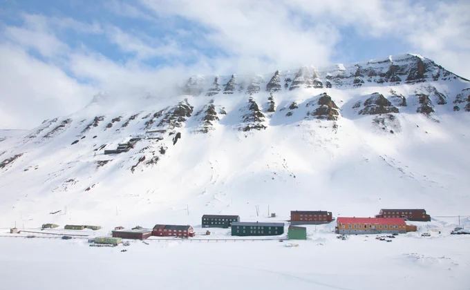 Lumipeitteisen vuoren rinteellä sijaitsee rakennuksia, ja vuoren huippu on osittain pilvien peitossa. Kuva: Marcel Schütz / Svalbard Photography.