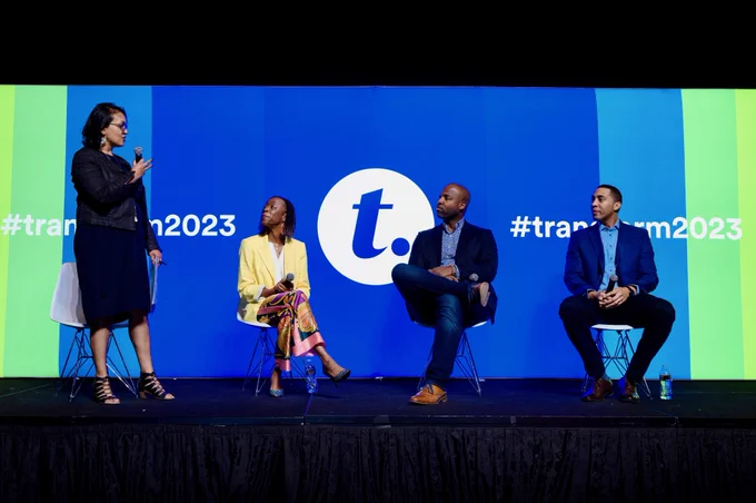 Meghna Majmudar speaks to three panelists on stage in front of a multicolored Transform 2023 background. 