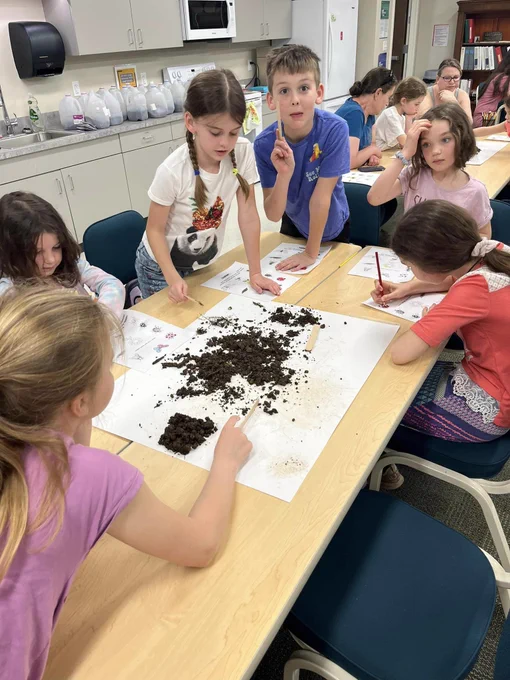 children at a table with worksheets and a pile of dirt. One child look directly at camera with a surprised expression.