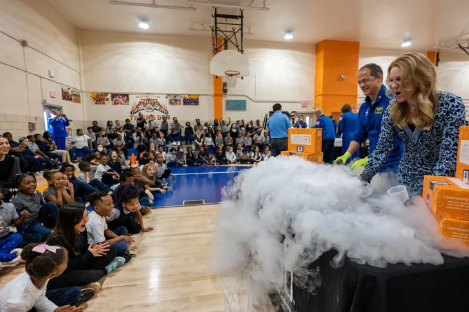 NASA astronaut Tom Marshburn and an educator conduct a science demonstration at Amidon-Bowen Elementary School in Washington, DC. Marshburn and the educator are standing on the far right of the photo, behind a black table with clouds of white smoke billowing off of it. The experiment is taking place in a school gymnasium, with several rows of excited-looking children in front of and to the side of the table. Credit: NASA/Keegan Barber