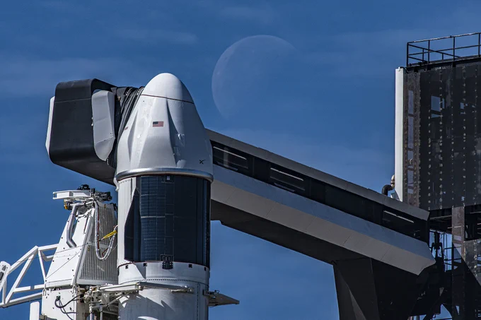 A SpaceX Dragon spacecraft, a conical white capsule, perches atop a Falcon 9 rocket. The launch tower is on the far right of the image, with a launch arm extending across the photo to connect behind Dragon. In the background, the faint outline of a large Moon can be seen against a clear blue sky. Credit: SpaceX