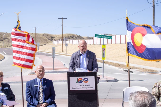 Shane Ferguson, director of region 2 for the Colorado Department of Transportation, speaks at a podium with the American and Colorado flags blowing in the wind behind him.
