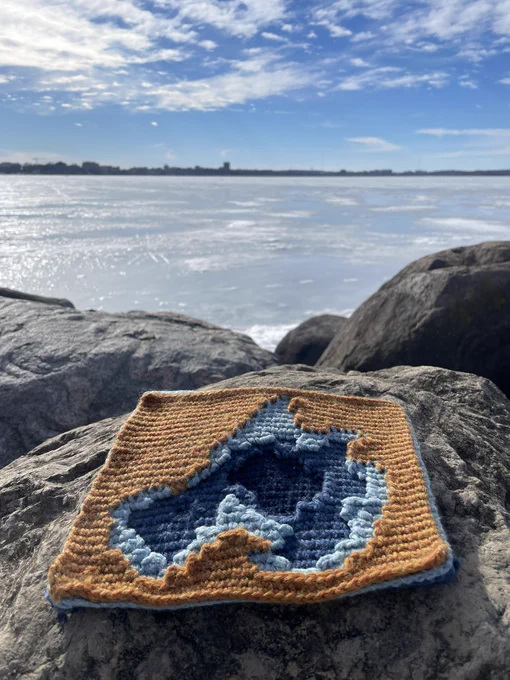 A crocheted map of Lake Mendota sitting on a rock overlooking a frozen Lake Mendota.