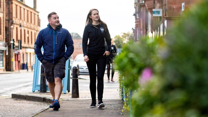A man and women in dark outdoor active wear look to their left as they enjoy a walk along the street of a city. 