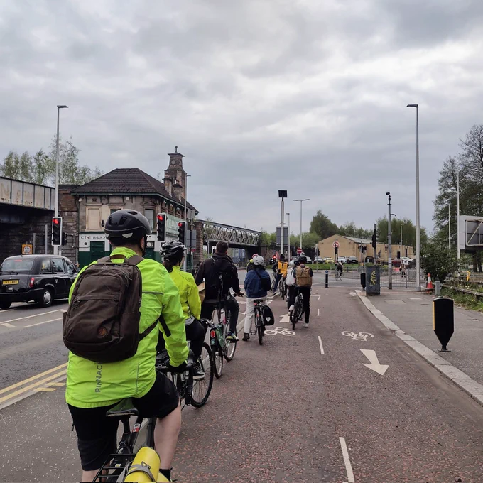 Picture showing a queue of roughly a dozen people on bikes, waiting to cross at traffic lights on the South City Way cycle route, on the southside of Glasgow.