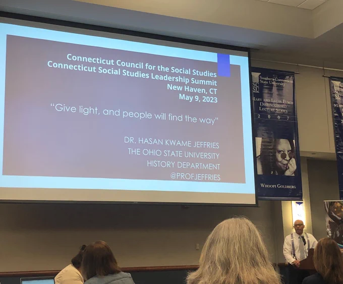 Photograph of a presentation projected on a white screen. There is a Black man, Dr. Hasan Kwame Jeffries, standing at a podium and the backs of people's heads. 