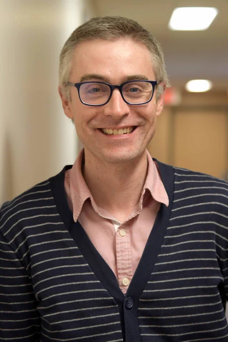 Headshot of Dr. Ryan MacNiel from the mid-chest up. Dr. MacNeil stands in a hallway, smiling while he leans against the wall on his left.