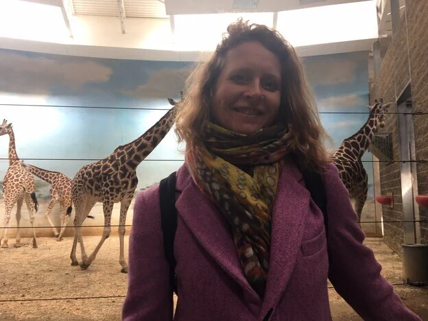 Sarah Riggs smiling to the camera wearing a purple jacket and patterned scarf, standing in front of an enclosure with giraffes in it