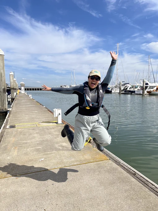 This photo shows Jessika (a filipino woman) in a wetsuit, dry pants, boots, a life jacket, and a green cap jumping into the air on a marina dock in San Francisco. Blue skies and boats can be seen in the background.