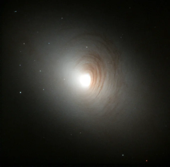 The bright, pale center of a galaxy shines against the black of space. Brown rings of dust encircle it, and small bright points of light, which are star clusters, are seen near the galaxy.