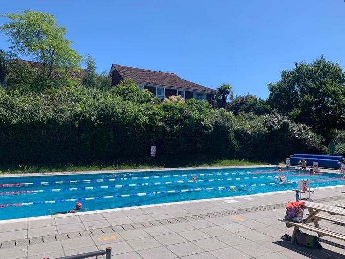 An outdoor swimming pool with lanes. It is positioned next to some high hedges.