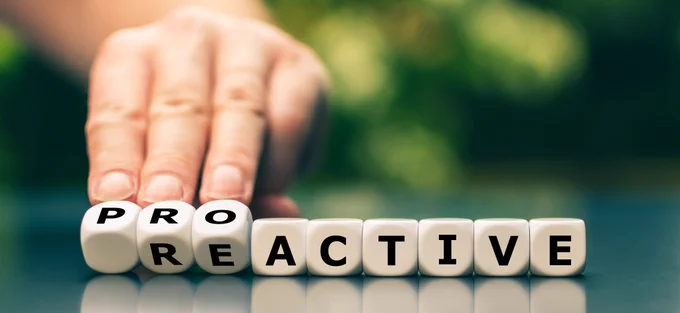 A row of 9 dice spells out the word "reactive" with one letter displayed on each die. In the background, a hand reaches to roll the "re" to "pro" to spell "proactive." The image represents a shift from "reactive" to "proactive."