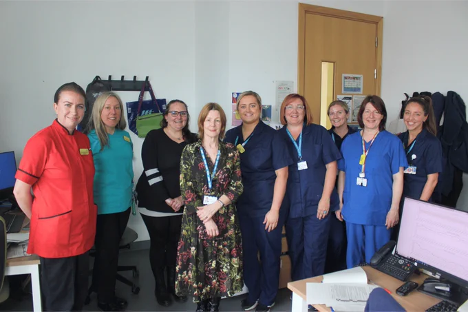Maria is pictured smiling with a group of eight NBT staff members who are wearing scrubs and clinical uniforms.