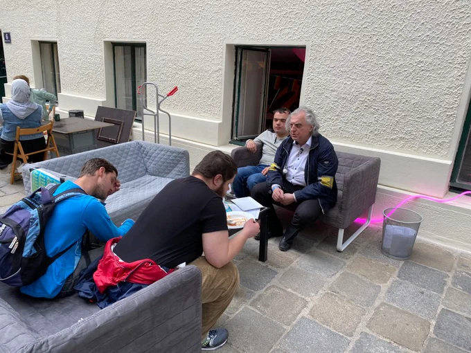 Vier Menschen auf der Straße sitzen auf zwei Sofas. Den Hintergrund bildet eine Fassade mit Fenstern, eines ist geöffnet.