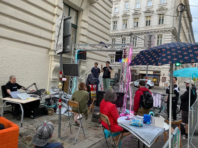 Auf einer mit 4-Punkt-Traversen eingerahmte Bühne stehen zwei Menschen hinter einem Mikrofon, einer gebärdet. Sieben Menschen sitzen oder stehen Richtung Bühne, eine weitere sitzt vor der Bühne, Richtung Publikum. Im Hintergrund befinden sich Häuserfassaden, Absperrgitter und parkende Autos. Ein Verkehrsschild zeigt das Ende der Geschwindigkeitsbegrenzung auf 30 km/h an. Zwei Regenschirme sind wie Sonnenschirme aufgestellt. Auf einem Tisch befindet sich ein Mischpult, ein Mikrofon und zwei Laptops.