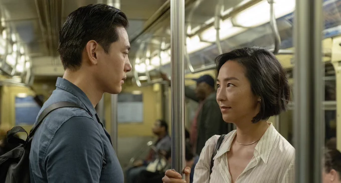 A film still from Past Lives. Teo Yoo and Greta Lee are in a subway, holding onto a pole, facing each other and looking into each others eyes sincerely. Teo Yoo wears a blue shirt and backpack. Greta Lee wears a white-striped shirt and a bag on her right shoulder. 