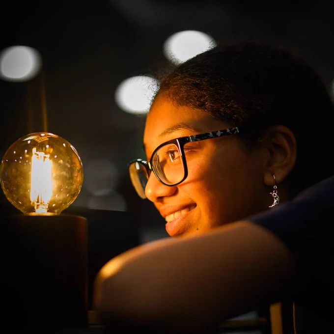 Close up of your female side profile wearing glasses as she smiles and looks into a light bulb within a dark room.