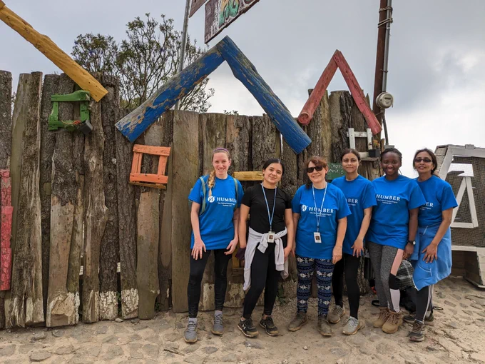 A group of six people stand in front of a wooden structure. Five of them are wearing Humber College t-shirts.