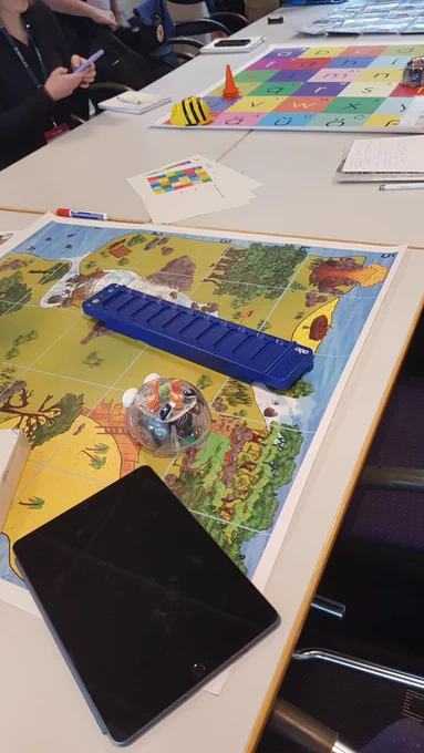 Eine Schatzkartenmazte und eine Buchstabenmatte liegen auf Tischen, darauf verteilt verschiedene Blue-Bot und Bee-Bot Lernroboter, Arbeitszettel, eine blaue Programmierleiste, ein Tablet und Notizblöcke
