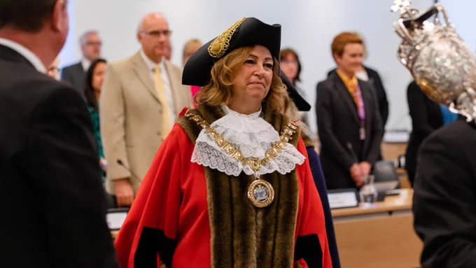 Cllr Linda Mascot, dressed in red ceremonial Mayor robes, with hat and large gold chain. She is standing in Chelmsford City Council's council chamber. 