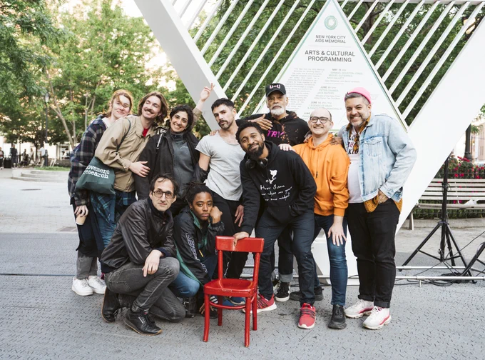 A group of people pose for a photo on a sunny day. They smile and huddle together at varying levels. They stand around a small red chair in the center of the image.