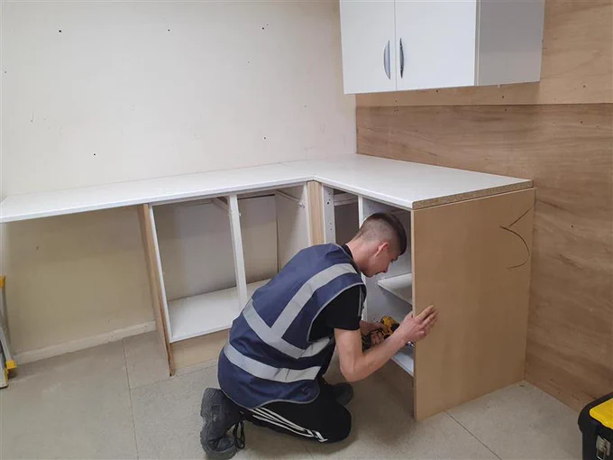 Student wearing a high vis vest drilling into a cupboard in a kitchen like setting  