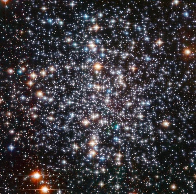 Thousands of bright points of light on a black background fill the field of view. They appear somewhat more concentrated near the centre of the image. They have a variety of colours, with the most prominent stars appearing blue or yellow-orange. The brighter stars also display four diffraction spikes.