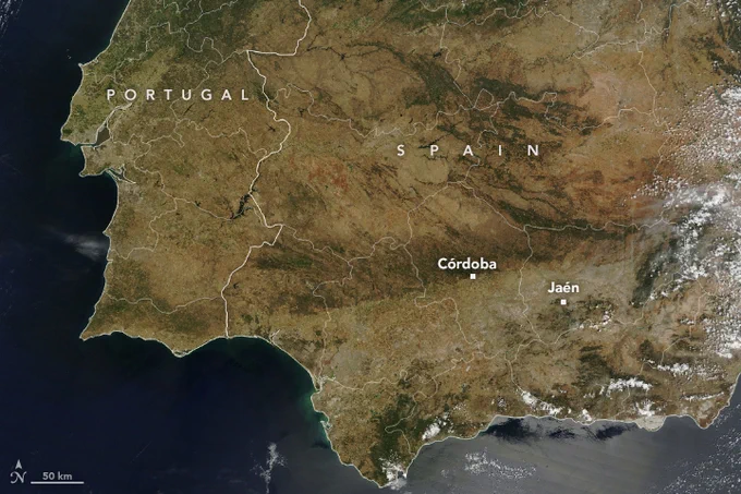 Satellite image of Spain from May 2023, during a drought. The area is now mostly brown with patches of dark brown and grey where there was green vegetation a year ago. The ocean to the left and bottom of the region is dark blue, with some cloud coverage in the bottom right.