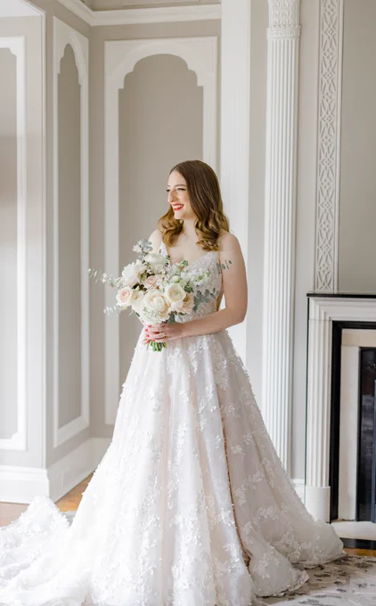 I'm standing in my wedding dress holding my pink/white/green bouquet in front of a fireplace with light shining in on me. I'm smiling because it's so special, and the photo has a light and airy feel to it.