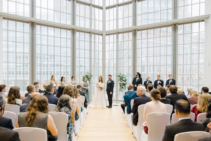 The wedding ceremony. We're pausing for people to take pictures before starting. The photo was taken from the end of the aisle, and you can see us smiling at all our friends and family. Behind us is a wall of windows that overlooks Elgin street in Ottawa, it's letting in a lot of natural light.