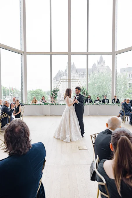 A photo of our first dance, we're arm in arm. Behind us is the head table with our friends and the hexagonal arch from the ceremony room. Behind it is giant windows, and we're overlooking the Chateau Laurier.
