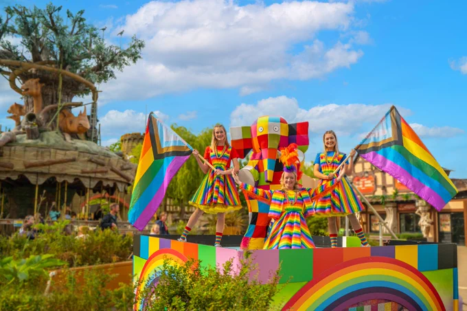 Three members of Elmer's Colour Krewe with Elmer the Patchwork Elephant on their stage in Adventure Point, next to the Adventure Tree Carousel. They are wearing bright rainbow outfits and holding the Progress Pride flag.