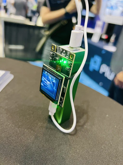 A tiny screen mounted on an ESP32 camera module, attached to a green power bank on a table. The screen shows Alex's hand with a green box around it.