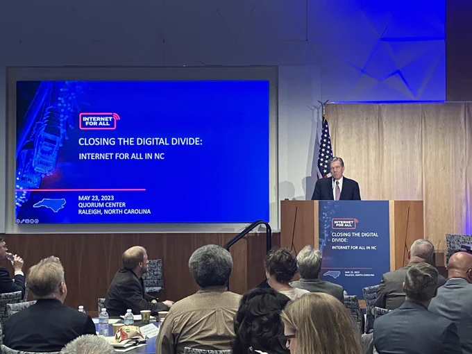 North Carolina Governor Roy Cooper pictured speaking at a podium with a slide displaying: "Closing the Digital Divide: Internet for All in NC" to his left. 