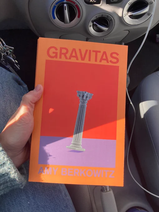 my hand holding the colorful US copy of Gravitas which shows a Corinthian column starting to tip over. I am in my car and the air conditioning knobs are visible behind the book.