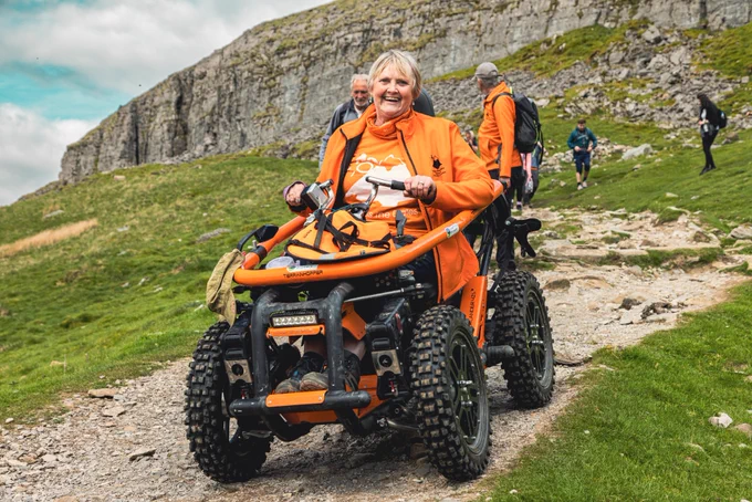 Debbie North is her orange all-terrain wheelchair coming down from the path at Pen-y-ghent. It is a very happy  photograph.