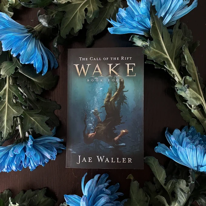 Photo of a paperback book. The book title is The Call of the Rift: Wake. The cover is an illustration of a young woman sinking upside-down through the ocean, wearing a glowing golden ring on her finger. The book itself is surrounded by large blue flowers and green leaves to match the cover's colour palette.