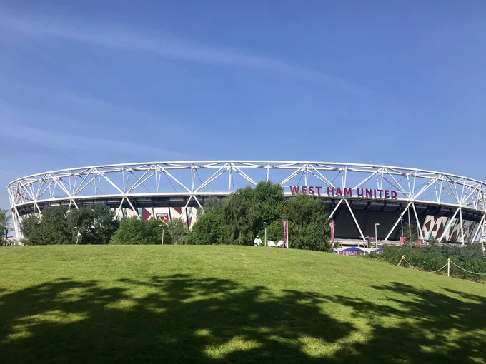 London Stadium with a sunny grassy bank with the shadows of trees on it in the foreground