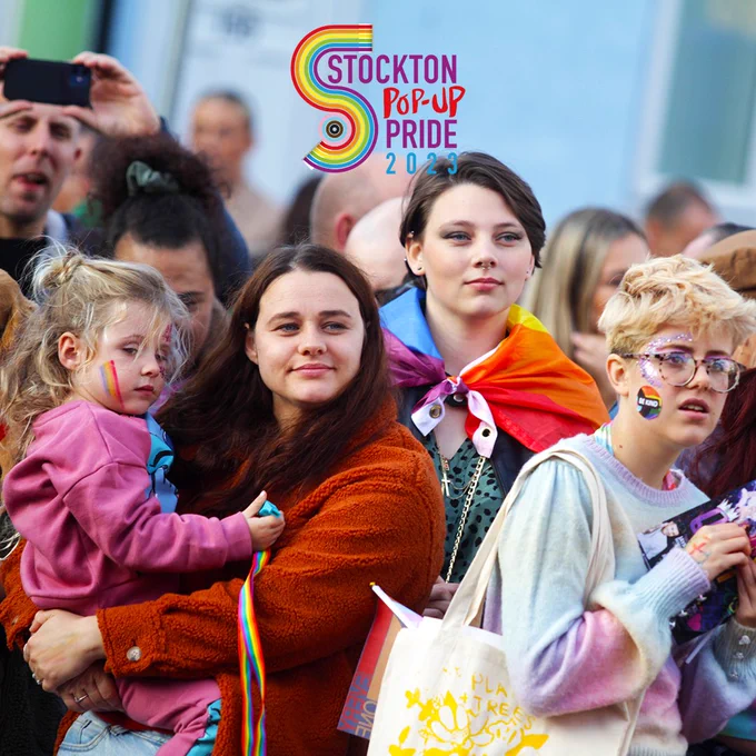 A crowd of people outside. In the foreground is four people. On the left is an adult holding a child with rainbows painted on their cheeks. To the right of them is a young person with short brown hair and a pride flag wrapped around their shoulders. On the right is a young person with short blonde hair and glasses with a pride flag painted on their cheek and a tote  bag on their shoulder.