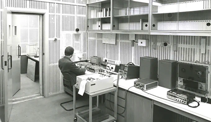 A man wearing a suit sits at a desk in a very 70s recording studio, he is surrounded by vintage equipment, including reel to reel recorders