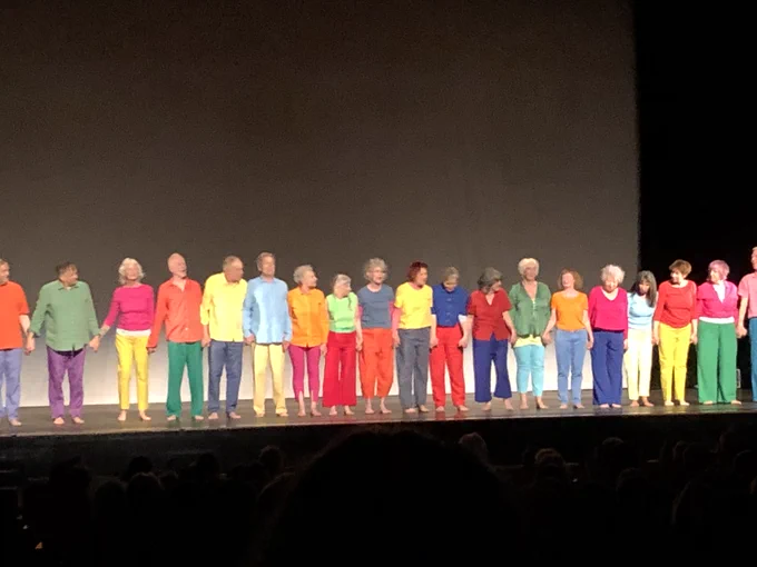 A long line of dancers of all genders stands along the front of a plain grey and black stage, holding hands. They are all wearing bright primary coloured shirts and trousers.