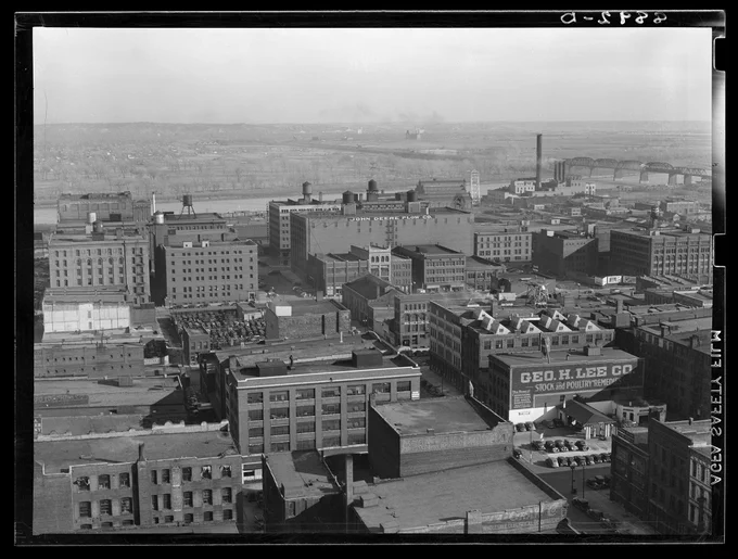 Historic black and white photo taken from a high angle of a city downtown area sitting beside a river. The city buildings are largely made of brick.