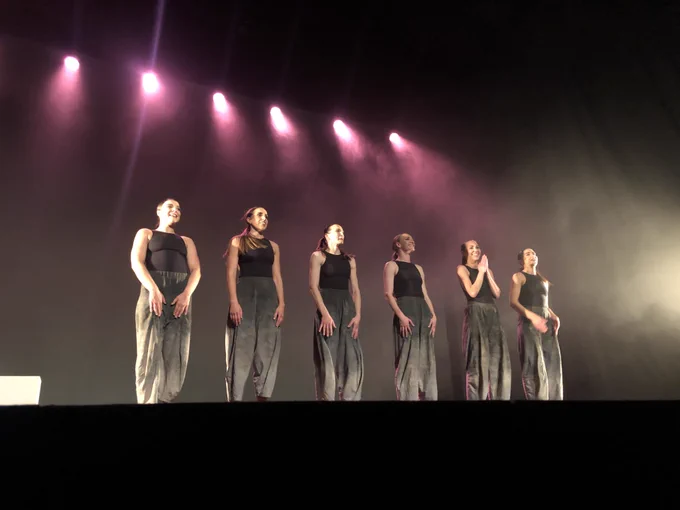A line of 6 dancers, all wearing matching dark baggy trousers and black tops standing before taking a bow onstage in a dark theatre space with pink lighting and text. They are all smiling at the audience.