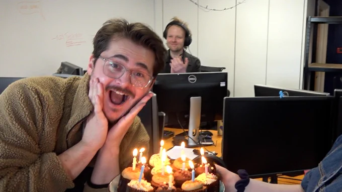 Tom looks overjoyed leaning over a plate of cupcakes with candles lit in them. Alex claps in the background. 