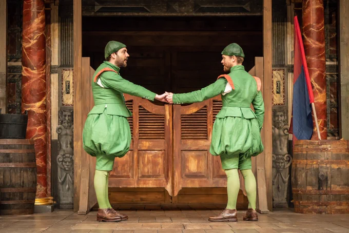 Two performers in identical green outfits stand sideways with one arm outstretched towards each other holding hands. 

George Fouracres as Dromio of Ephesus and Jordan Metcalfe as Dromio of Syracuse in The Comedy Of Errors (2023).