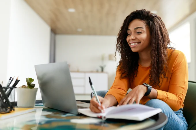 A woman looks at a laptop screen and writes in a notebook